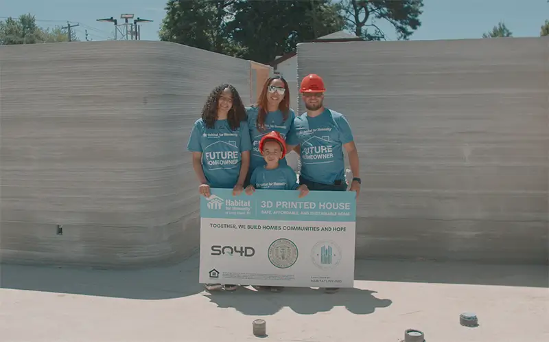 A family holding up a Habitat for Humanity sign in a 3D printed house